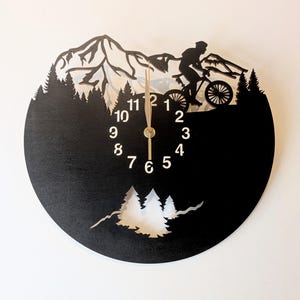 May include: Black wooden wall clock featuring a mountain biking scene. The clock face is black with white numerals and hands. A silhouette of a cyclist on a mountain bike is depicted, along with mountain and forest cutouts. The clock measures approximately 12 inches in diameter.
