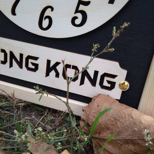 May include: A wooden clock with the word "HONG KONG" cut out below the numbers 4, 5, 6, and 7. The clock is set against a black background. A small plant and a brown leaf are in the foreground.
