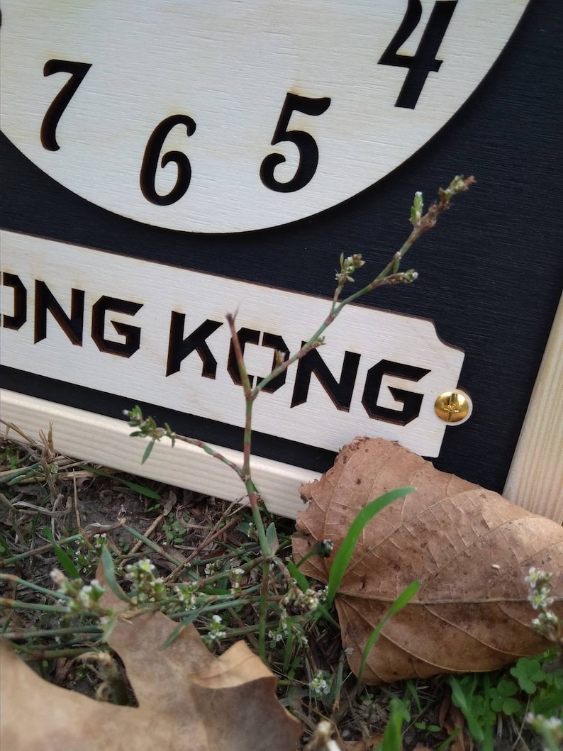 May include: A wooden clock with the word "HONG KONG" cut out below the numbers 4, 5, 6, and 7. The clock is set against a black background. A small plant and a brown leaf are in the foreground.