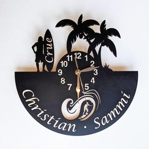 May include: A black wall clock with a semi-circular base featuring the names Christian and Sammi. The design includes a silhouette of a person with a surfboard labeled Crue, two palm trees, and a graphic of a surfer riding a wave with clock hands.