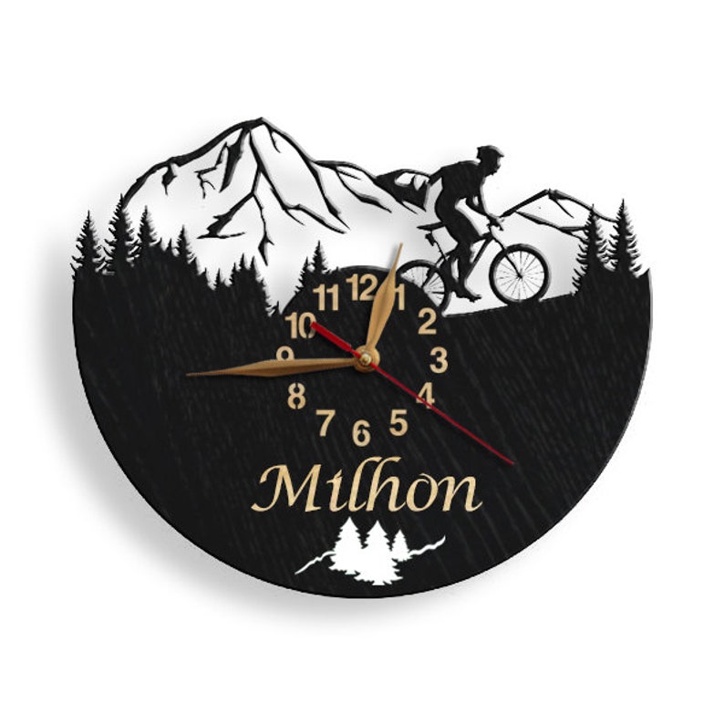 May include: Black, round wall clock with a mountain biking theme. The clock features a silhouette of a cyclist, mountains, and trees. The clock hands are gold, and the numbers are in a gold font. The name "Milhon" is displayed in a gold font.