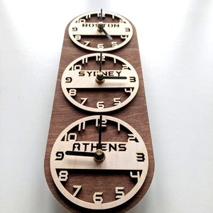 May include: A wooden multi-time zone clock with three circular faces. Each clock face displays a different city: Boston, Sydney, and Athens. The clock hands are black, and the numbers are cut out of the light-colored wood.