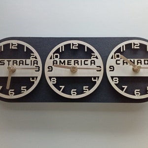 May include: A wooden world time zone clock with three circular faces. Each clock face is labeled with a different continent: Australia, America, and Canada. The clock hands are light brown, and the background is black.