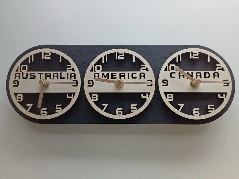 May include: A wooden world time zone clock with three circular faces. Each clock face is labeled with a different continent: Australia, America, and Canada. The clock hands are light brown, and the background is black.