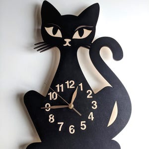 May include: A black cat-shaped wall clock with a light-colored face and hands. The clock features a stylized cat silhouette with a tail, ears, and eyes. The clock face displays the numbers 1 through 12.