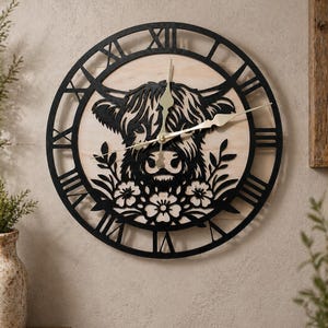 May include: A round wall clock featuring a black silhouette of a Highland cow's face and floral accents. The clock has a light-colored wooden background with black Roman numerals and hands. The clock is mounted on a neutral-toned wall.