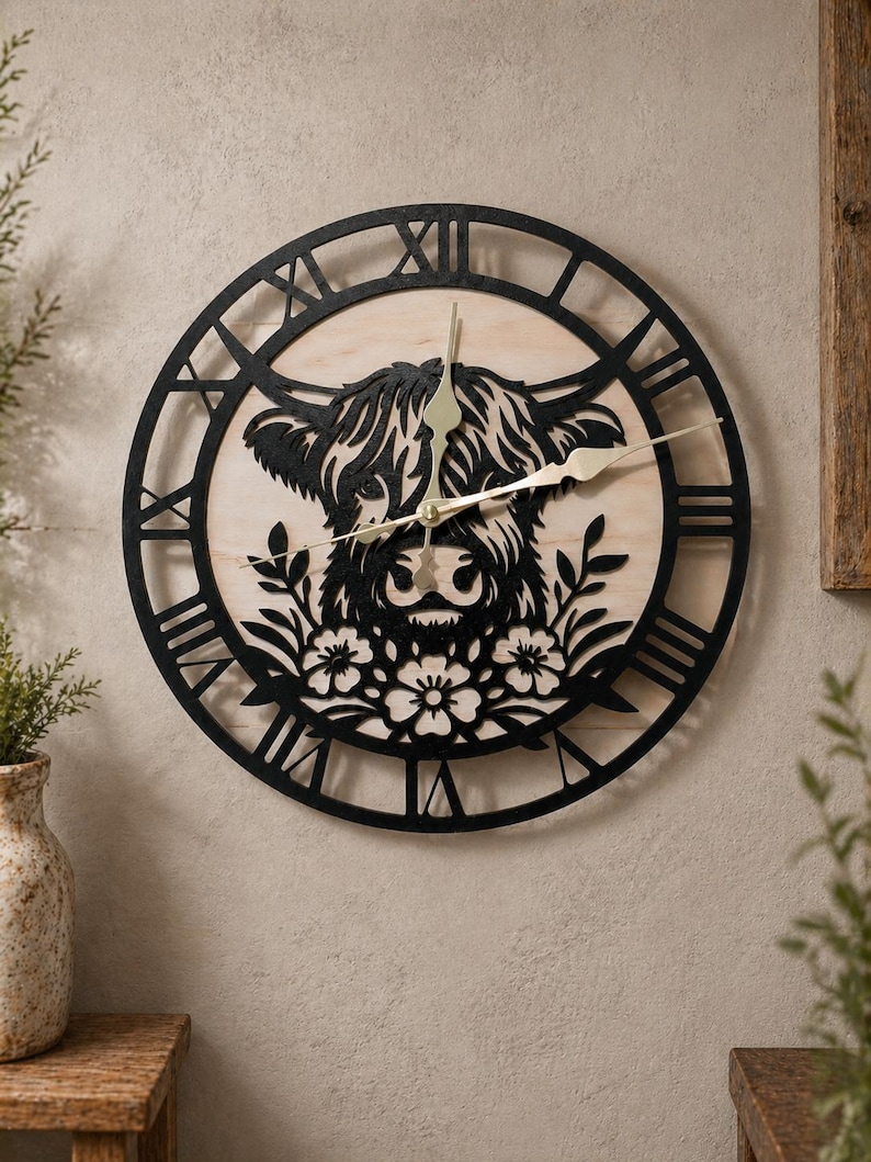 May include: A round wall clock featuring a black silhouette of a Highland cow's face and floral accents. The clock has a light-colored wooden background with black Roman numerals and hands. The clock is mounted on a neutral-toned wall.