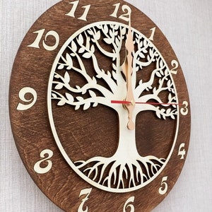 May include: A wooden wall clock featuring a tree of life design. The clock face is brown with white numbers and a white tree of life design in the centre. The clock hands are gold and red.