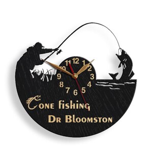 May include: Black wooden clock with a silhouette of a person fishing. The clock face has gold numbers and hands. The text "Gone fishing Dr Bloomston" is written below the clock face.