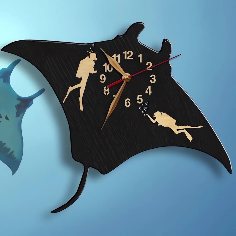 May include: Black wooden wall clock in the shape of a manta ray, featuring a clock face with gold hands and numbers. Two divers are depicted in silhouette. The clock is mounted on a blue wall, with a partial image of a blue manta ray.