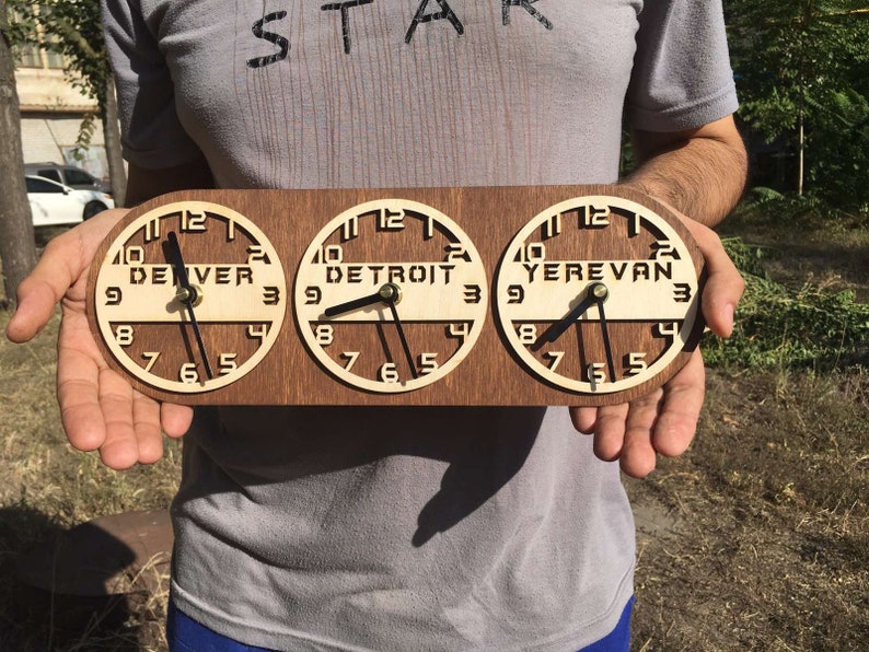 May include: A wooden clock with three circular faces, each displaying a different time zone: Denver, Detroit, and Yerevan. The clock hands are black, and the numbers are in a classic font. The clock is held by a person.