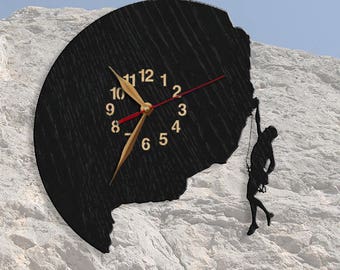 Puede incluir: Reloj de pared negro con números y manecillas doradas, con una silueta de escalador. El reloj tiene una superficie oscura y texturizada y un segundero rojo. El reloj está montado sobre una superficie rocosa blanca.