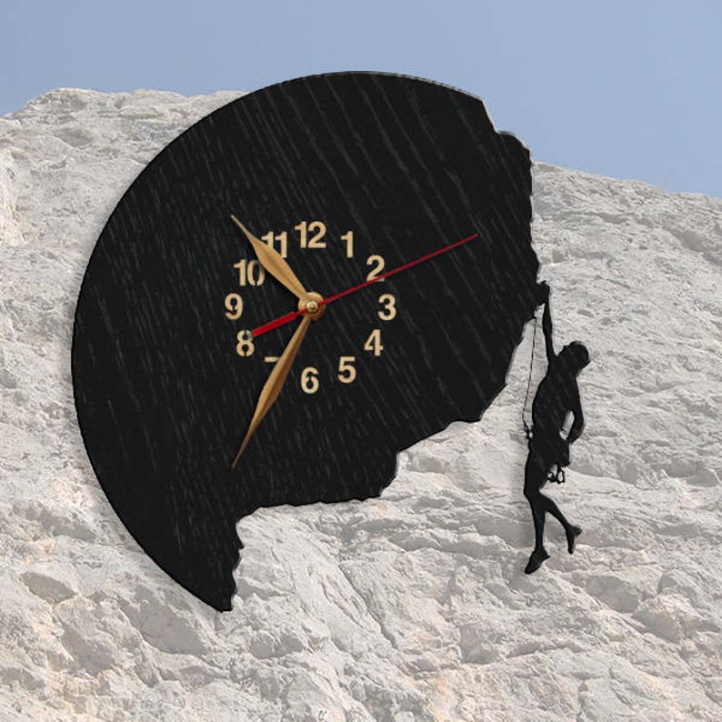 May include: Black wall clock with gold numerals and hands, featuring a climbing figure silhouette. The clock has a textured, dark surface and a red second hand. The clock is mounted on a white, rocky surface.