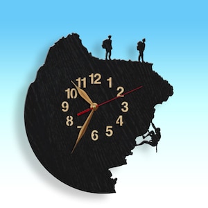Può includere: Orologio in legno nero a forma di montagna con scalatori in silhouette sul lato. Il quadrante dell'orologio ha lancette dorate e numeri dorati.