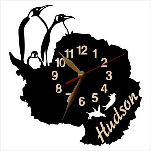 May include: Black and white silhouette of penguins and a clock face with gold hands and Roman numerals. The clock face is shaped like the continent of Antarctica. The text "Hudson" is written in a cursive font below the clock face.