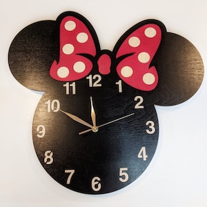 May include: Black clock with a red and white polka dot bow, shaped like Mickey Mouse's head. The clock face has white numbers 1 through 12 and gold hands.