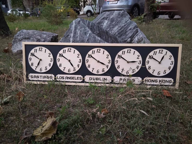 May include: A wooden world time clock with five clock faces, each displaying a different time. The clock faces are white with black numbers and hands. The names of the cities are printed below each clock face: Santiago, Los Angeles, Dublin, Paris, and Hong Kong.