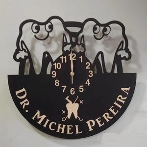 May include: Black and white clock with a dentist theme. The clock face is surrounded by two cartoon teeth with smiling faces. The clock face has gold numbers and hands. The clock is mounted on a black circular plaque with the text "Dr. Michel Pereira" in gold lettering.