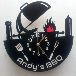 May include: Black and white clock with a grill design. The clock face has a black background with white numbers and hands. The grill design includes a grill with a lid, a flame, a spatula, a fork, and a bottle of BBQ sauce. The clock face also has the text "Andy's BBQ".