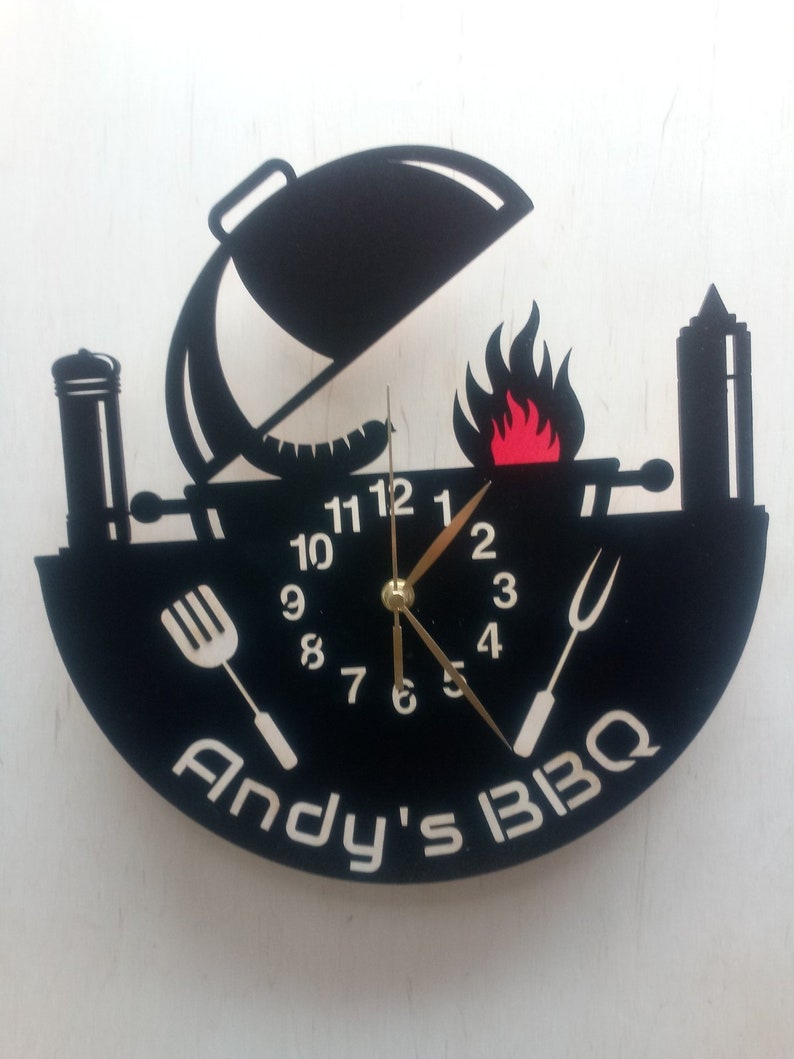 May include: Black and white clock with a grill design. The clock face has a black background with white numbers and hands. The grill design includes a grill with a lid, a flame, a spatula, a fork, and a bottle of BBQ sauce. The clock face also has the text "Andy's BBQ".