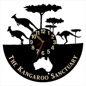 May include: Black and white clock with a silhouette of kangaroos and trees. The clock face has a black background with white numbers and gold hands. The clock is surrounded by a black circle with the text "The Kangaroo Sanctuary" in white.