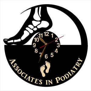 May include: A black and tan wall clock with a podiatry theme. The clock features a silhouette of a foot skeleton, a clock face, and the words "Associates in Podiatry". The clock hands are gold.
