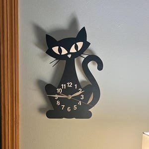 May include: Black cat-shaped wall clock with white eyes and a black tail. The clock face has black numbers and gold hands.