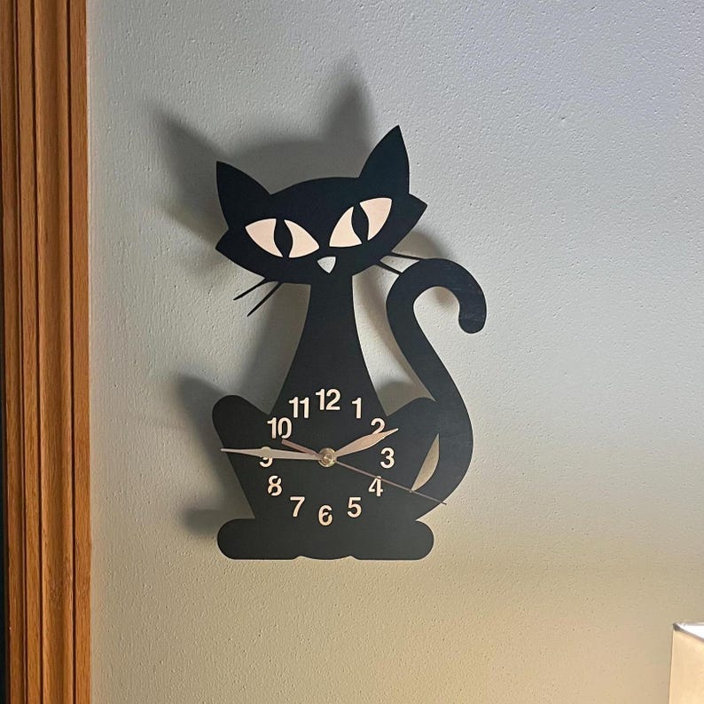 May include: Black cat-shaped wall clock with white eyes and a black tail. The clock face has black numbers and gold hands.