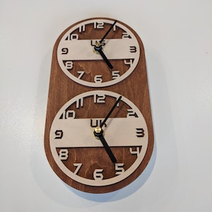 May include: A wooden wall clock with two circular clock faces. The clock faces have a light-colored interior with black numerals and hands. The clock is mounted on a dark brown wooden base. The text "UK" is visible on the clock face.