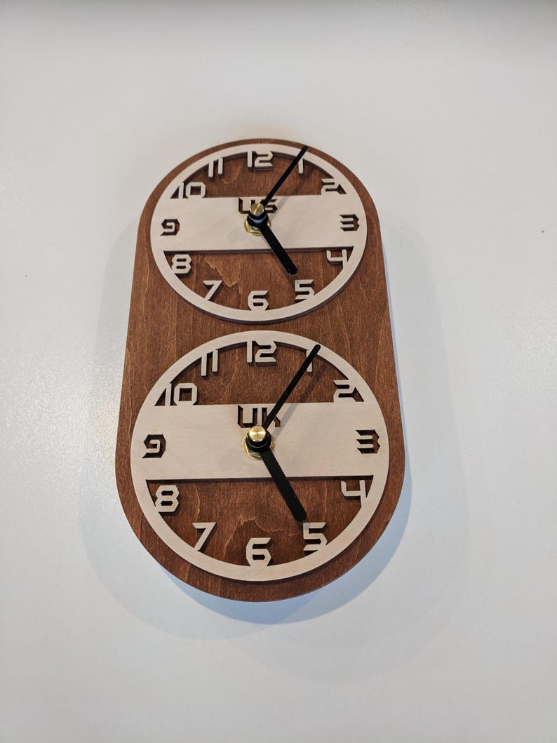 May include: A wooden wall clock with two circular clock faces. The clock faces have a light-colored interior with black numerals and hands. The clock is mounted on a dark brown wooden base. The text "UK" is visible on the clock face.