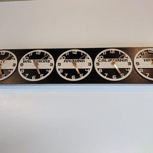 May include: A black wooden wall clock with five circular clock faces. Each clock face displays a different time zone: GMT, Baltimore, Arizona, California, and Hawaii. The clocks have white faces, black numbers, and gold hands.