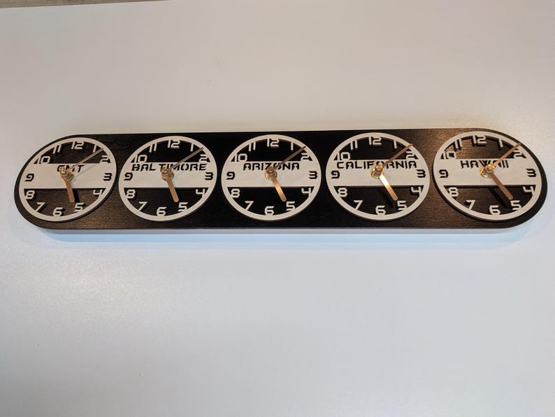 May include: A black wooden wall clock with five circular clock faces. Each clock face displays a different time zone: GMT, Baltimore, Arizona, California, and Hawaii. The clocks have white faces, black numbers, and gold hands.