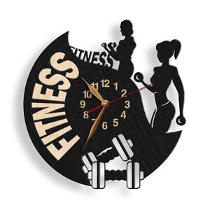 K&ouml;nnte beinhalten: Schwarze und wei&szlig;e Wanduhr mit Holzmaserung. Das Zifferblatt der Uhr zeigt eine Uhr mit r&ouml;mischen Ziffern und eine Silhouette von zwei Frauen, die Gewichte heben. Die Uhr ist von dem Wort "FITNESS" in Holzmaserung umgeben.
