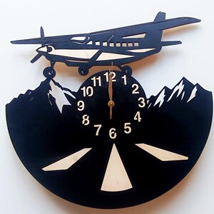 May include: Black vinyl record clock with a silhouette of a plane flying over mountains. The clock face has white numbers and a gold hand. The plane is made of wood and has a light brown color.