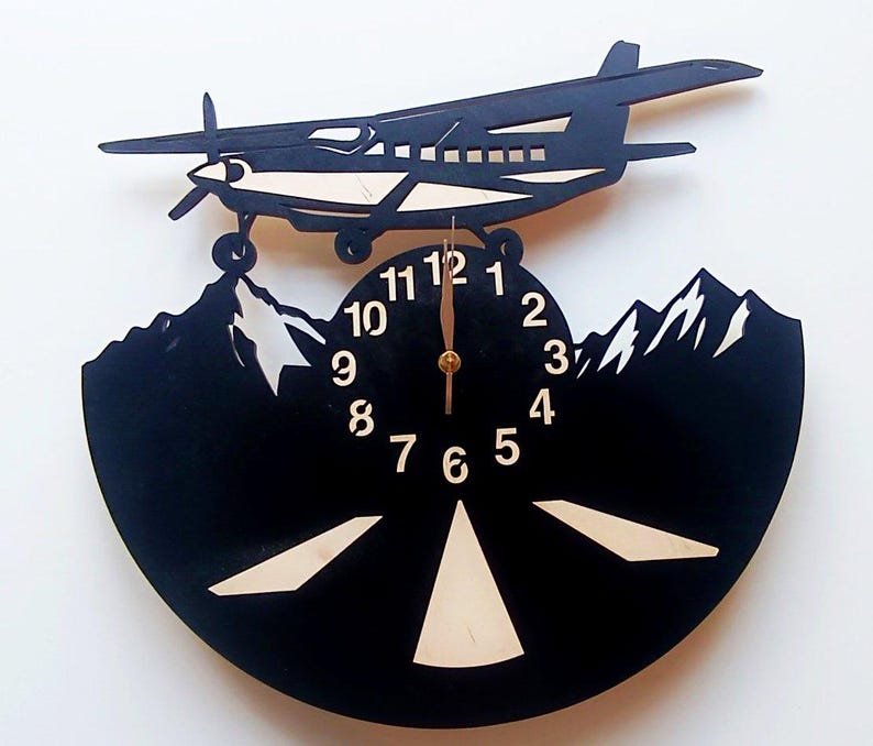 May include: Black vinyl record clock with a silhouette of a plane flying over mountains. The clock face has white numbers and a gold hand. The plane is made of wood and has a light brown color.