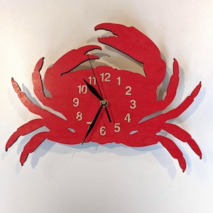 Op de afbeelding: Een rode houten wandklok in de vorm van een krab. De wijzerplaat is geïntegreerd in het lichaam van de krab, met zwarte wijzers en witte cijfers. De klauwen en poten van de krab steken naar buiten.