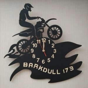 May include: Black metal wall clock featuring a silhouette of a motocross rider jumping over a flame. The clock face has gold numbers and hands. The clock is signed "Barkdull 173".