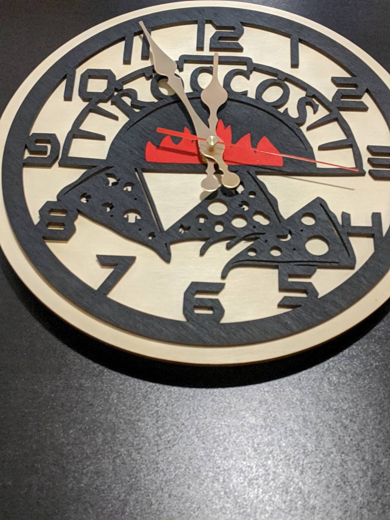 May include: A wooden clock with a pizza design. The clock face features a pizza with cheese and pepperoni, a red flame, and the word "Pizza" written around the edge. The clock hands are black and gold.