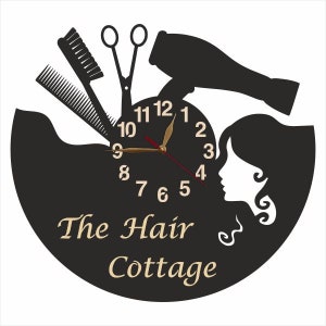 May include: Black and white clock with a hair salon theme. The clock face features a hair dryer, scissors, combs, and a woman's silhouette. The clock face reads "The Hair Cottage".