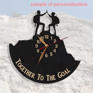 May include: Black wooden clock with a mountain silhouette and two figures high-fiving on top. The clock face has gold hands and Roman numerals. The text "TOGETHER TO THE GOAL" is written in a gold font on the bottom of the clock.