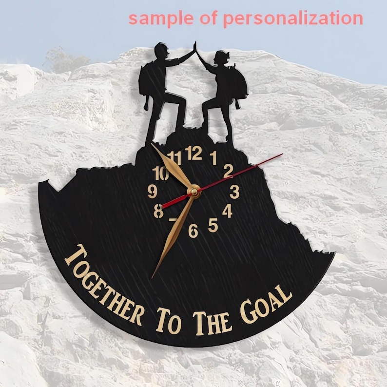 May include: Black wooden clock with a mountain silhouette and two figures high-fiving on top. The clock face has gold hands and Roman numerals. The text "TOGETHER TO THE GOAL" is written in a gold font on the bottom of the clock.
