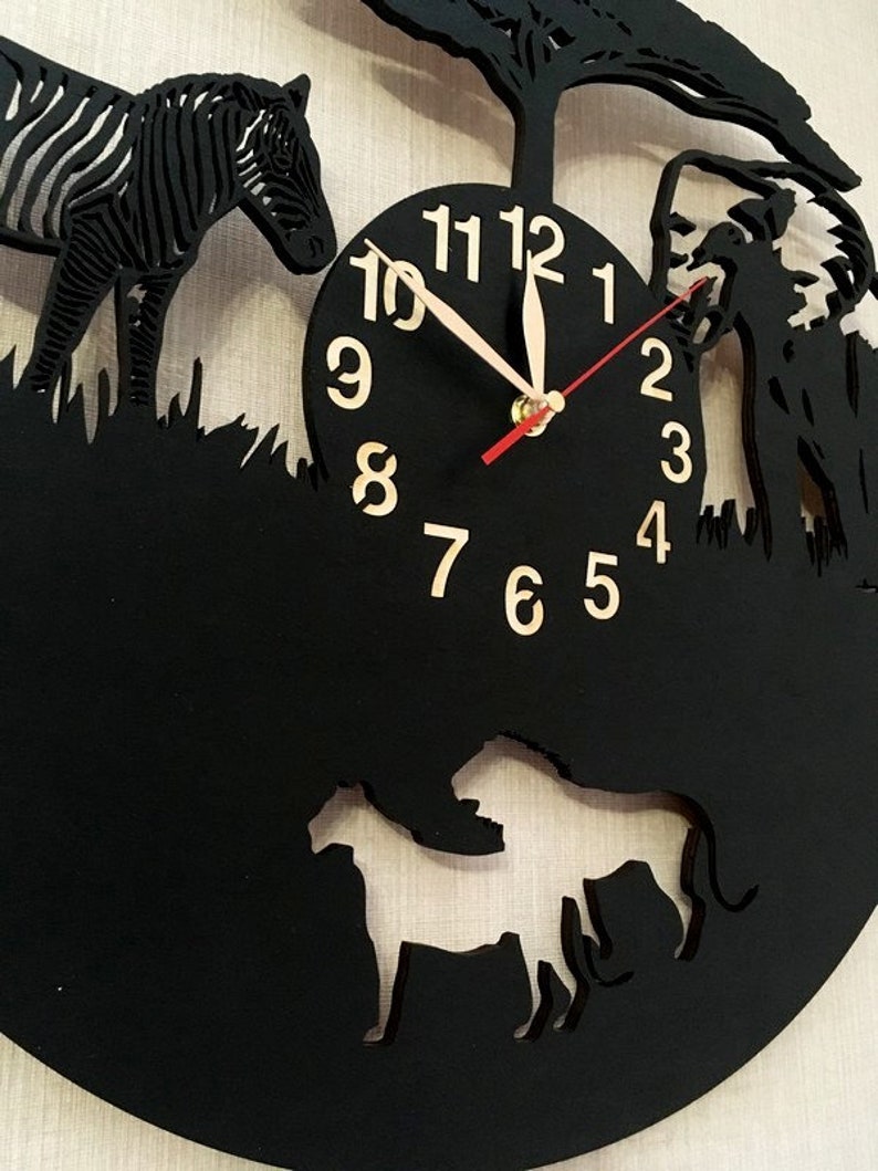 May include: Black clock with a white background and gold numbers. The clock face is surrounded by cutouts of a zebra, a lion, and a tree. The clock hands are red and white.