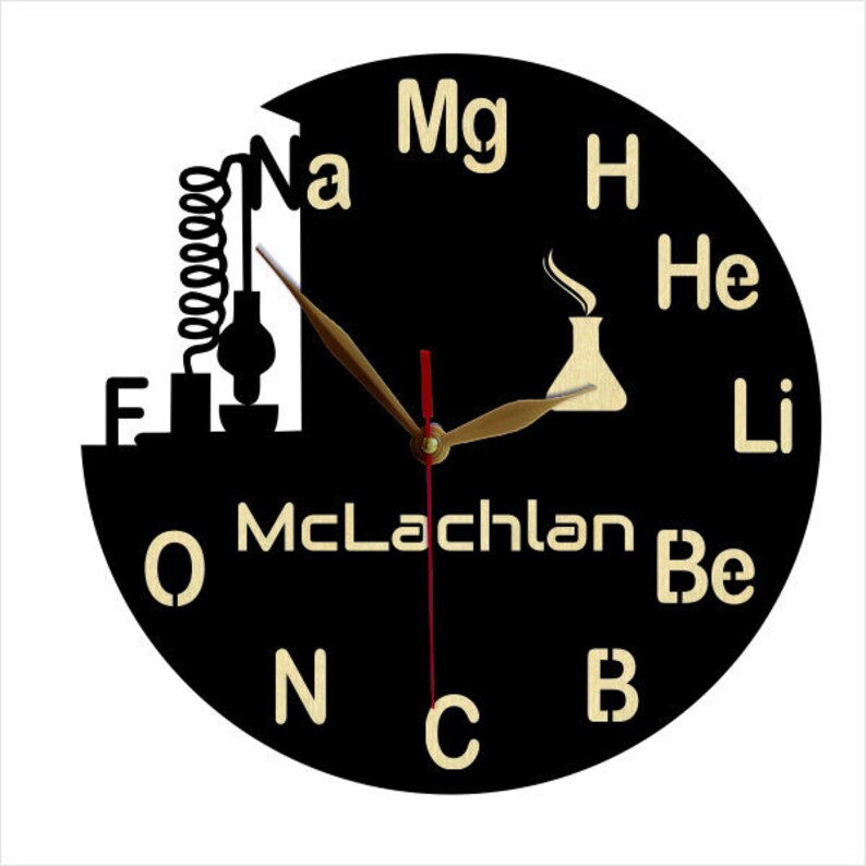 May include: A black round clock with gold hands and gold lettering. The clock face features the chemical symbols for various elements, including Mg, H, He, Li, Be, O, N, C, and Na. The clock also features a small illustration of a beaker and a coiled tube.
