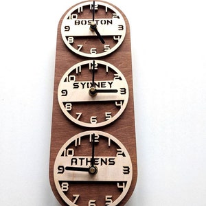 May include: A wooden wall clock displaying the time in Boston, Sydney, and Athens. The clock has three circular faces with the city names in bold letters. The clock hands are black, and the wood has a warm brown tone.
