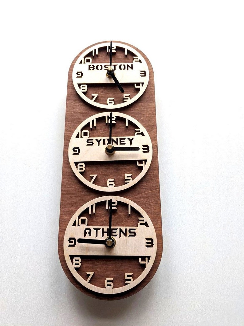 May include: A wooden wall clock displaying the time in Boston, Sydney, and Athens. The clock has three circular faces with the city names in bold letters. The clock hands are black, and the wood has a warm brown tone.