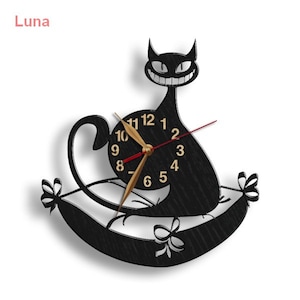 May include: Black cat wall clock with a clock face that reads 10, 11, 12, 1, 2, 3, 4, 5, 6, 7, 8, 9. The cat is sitting on a pillow with a bow on each side. The clock is titled "Luna".