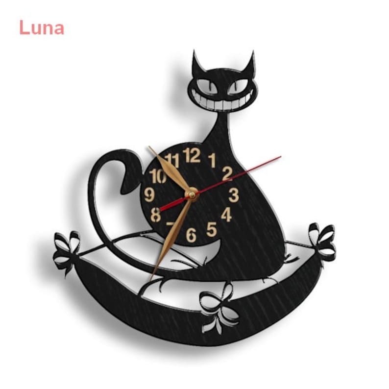 May include: Black cat wall clock with a clock face that reads 10, 11, 12, 1, 2, 3, 4, 5, 6, 7, 8, 9. The cat is sitting on a pillow with a bow on each side. The clock is titled "Luna".