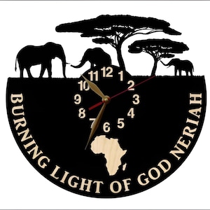 Puede incluir: Reloj de vinilo negro con una silueta de elefantes y &aacute;rboles. La esfera del reloj tiene una incrustaci&oacute;n de madera del continente africano. El texto "BURNING LIGHT OF GOD NERIAH" est&aacute; impreso alrededor de la esfera del reloj.