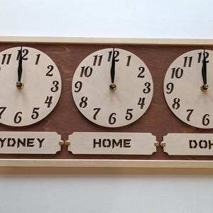 May include: Wooden world time clock with three circular clock faces. Each clock face has black numerals and hands. The clock displays the time for Sydney, Home, and Doha. The frame is light brown.