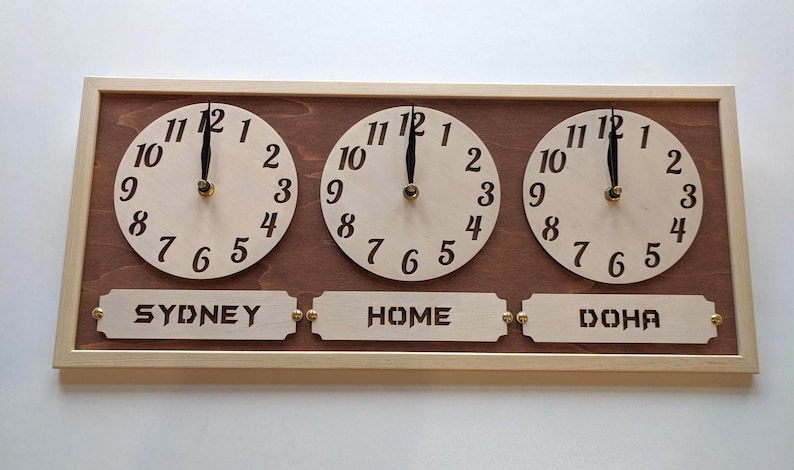 May include: Wooden world time clock with three circular clock faces. Each clock face has black numerals and hands. The clock displays the time for Sydney, Home, and Doha. The frame is light brown.